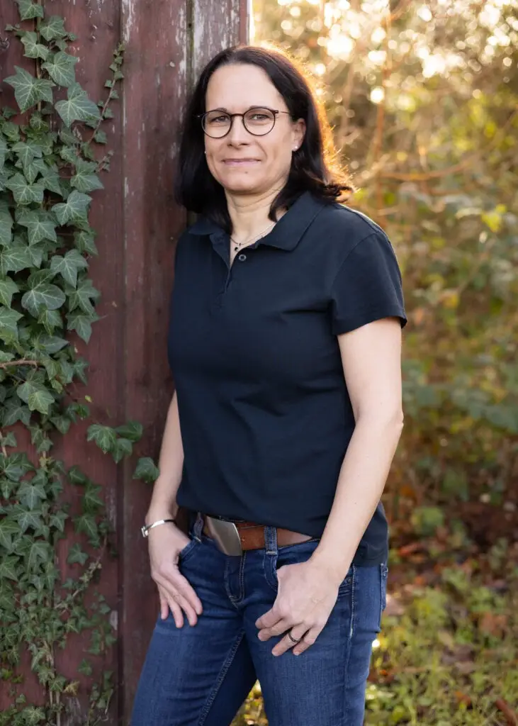 Frau mit dunklen Haaren und dunkler Brille, an einem Holztor lehnend mit schwarzem Polo und Jeans